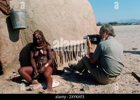 Ein Mann filmt eine Frau, die in ihrem Dorf in Namibia sitzt. Stockfoto