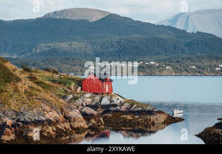 Malerische norwegische Fischerhütte, bekannt als „Hytte“, hoch auf dem felsigen Ufer mit Blick auf den ruhigen Fjord. Das kleine Boot ruht in ruhigem Wasser mit üppigem Berg Stockfoto