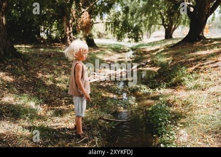 Kleines blondes Kind am Bach im Sommer mit Stöcken Stockfoto