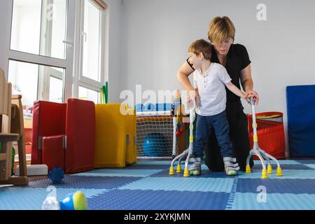 Der süße Junge mit Zerebralparese arbeitet mit einem Therapeuten an der körperlichen Rehabilitation mit den bariatrischen 4-Punkt-Stangen im Therapiezentrum Stockfoto