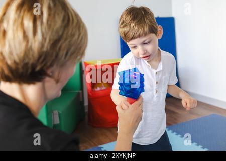 Der Physiotherapeut hilft einem kleinen Jungen mit Zerebralparese während einer spielerischen Rehabilitationssitzung, indem er farbenfrohe Werkzeuge verwendet, um Bewegung zu fördern Stockfoto