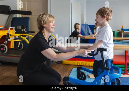 Niedlicher kleiner Junge mit Bewegungsstörungen steht mit einem Walker, während er mit einem Physiotherapeuten in einer unterstützenden Therapieumgebung arbeitet Stockfoto