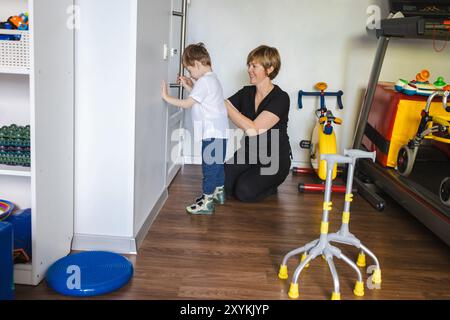 Ein engagierter Therapeut hilft einem kleinen Jungen mit Zerebralparese, sich in einer unterstützenden Therapieumgebung mit seiner Umgebung zu beschäftigen Stockfoto