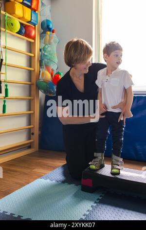 Ein engagierter Physiotherapeut hilft einem kleinen Jungen mit Zerebralparese, während er während seiner Rehabilitationssitzung auf der Reha-Plattform steht, um sein Gleichgewicht zu verbessern Stockfoto