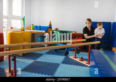 Der Junge wird von Therapeuten bei körperlichen Rehabilitationsübungen in hellem, spielerischem Therapieumfeld unterstützt. Handläufe zur Wiederherstellung der Gehfähigkeit Stockfoto