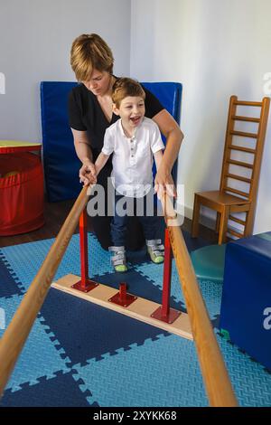 Junge männliche Kinder nehmen an einer Physiotherapie Teil, unterstützt von einer Betreuungsperson, und verwenden Parallelstangen, um die Mobilität zu verbessern. Handläufe zum Wiederherstellen von Laufschuhen Stockfoto