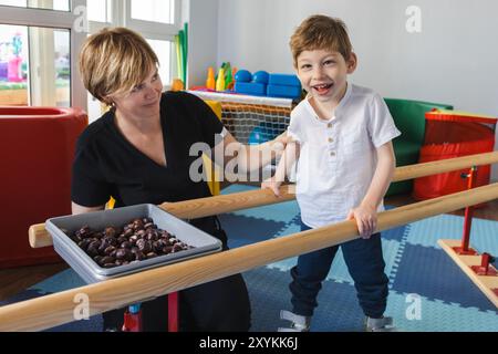 Junge mit Bewegungsstörungen lächelt, während er an der körperlichen Rehabilitation teilnimmt, unterstützt von einem Therapeuten, der in lebendiger R neben Handläufen steht Stockfoto