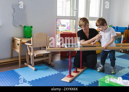 Junger süßer Junge mit Zerebralparese prahlt und balanciert mit Handläufen mit Hilfe des Physiotherapeuten in einem bunten Therapieraum Stockfoto