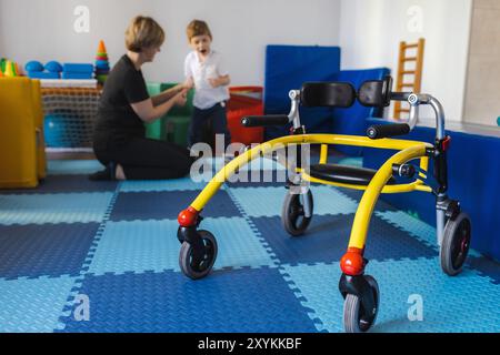 Junge mit Zerebralparese nimmt an der Physiotherapie Teil, unterstützt von einem Therapeuten in einer lebendigen Rehabilitationsumgebung mit Freirahmen-Walker Stockfoto