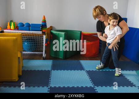 Lächelndes Kind mit Zerebralparese betreibt körperliche Rehabilitationsaktivitäten mit Physiotherapeuten in farbenfroher und unterstützender Umgebung Stockfoto