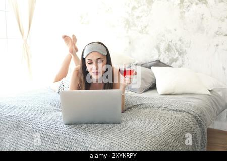 Fröhliche brünette Frau mit Glas Wein tragen schlafende Maske im Bett. Lesen und Schauen am Laptop Stockfoto