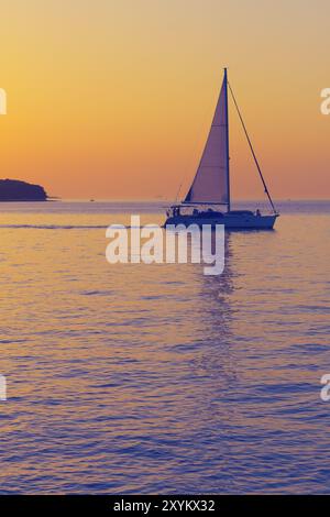 Das Segelboot verlässt die kroatische Küste nach Sonnenuntergang. Romantische Szene mit einem Silhouettenboot, das vor einem lebhaften, farbenfrohen Sonnenuntergang in Kroatien auf das offene Meer segelt Stockfoto