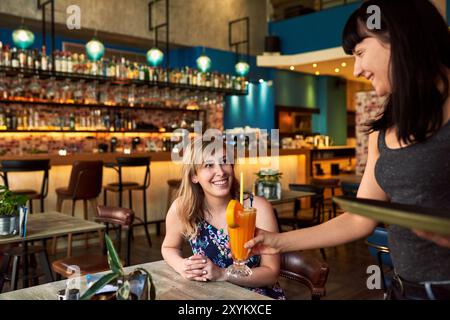 Gäste, Kellnerin oder servieren Getränke im Pub für Fruchtcocktail am Oktoberfest am Abend. Frauen, Lächeln oder Barkeeper Service mit Alkohol Glas auf Bestellung Stockfoto