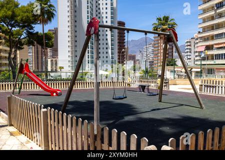 Foto eines leeren Kinderspielplatzes mit Schaukeln, Rutsche und einer Seesäge im Park, der von hohen Wohnblocks in der Stadt Be umgeben ist Stockfoto