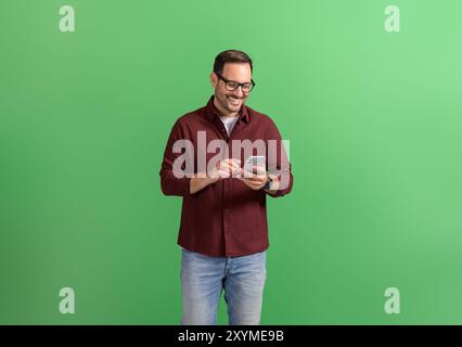 Junger gutaussehender Unternehmer mit Brille, der über Smartphone auf grünem Hintergrund Telekommutiert Stockfoto
