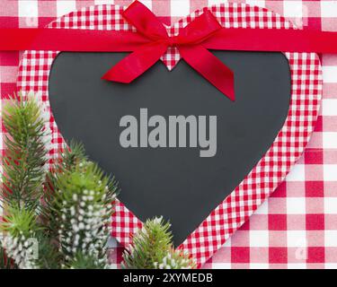 Weihnachtskartenrohling in Herzform mit Schleife und Ast auf roter Gingham-Tischdecke Stockfoto