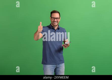 Positiver junger Geschäftsmann mit Smartphone, das das Daumen-hoch-Schild zeigt, während er auf grünem Hintergrund posiert Stockfoto