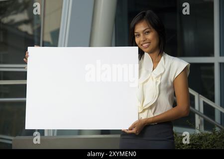 Eine fröhliche, schöne junge Geschäftsfrau hält ein leeres Schild in horizontaler Position, um außerhalb eines Büroparks Text einzufügen. 20s weiblich AS Stockfoto