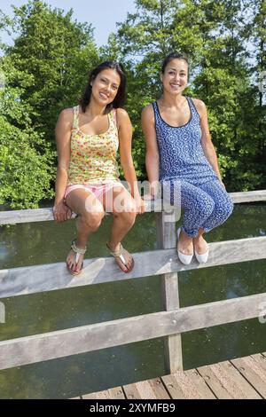 Zwei junge Frauen sitzen zusammen auf der Brücke Geländer oben Teich Stockfoto