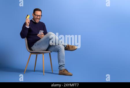 Aufgeregter junger Mann mit Kreditkarte und Handy, der glücklich auf Stuhl über blauem Hintergrund schreit Stockfoto