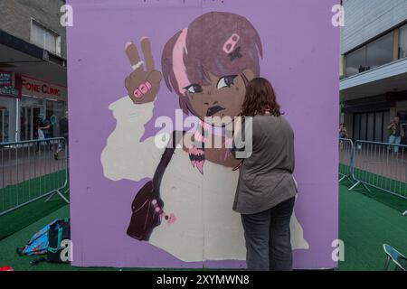 Southend on Sea, Großbritannien. 30. August 2024. Ein Künstler malt ein großes Wandgemälde im Cartoon-Stil an einer lila Wand in Southend-on-Sea. Die Figur hat rosa und braune Haare, einen gestreiften Schal und eine Friedenszeichen-Geste mit rosa Nägeln und einem bandagierten Finger. Die Szene ist Teil einer öffentlichen Kunstveranstaltung mit Metallbarrieren und Zuschauern im Hintergrund, die die Lebendigkeit des zeitgenössischen urbanen Ausdrucks einfangen. City Jam wird offiziell eröffnet und über 200 Künstler aus aller Welt malen Wandbilder. Penelope Barritt/Alamy Live News Stockfoto
