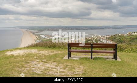 Eine Bank am South West Coast Path mit Blick in Richtung Fortuneswell und Chesil Beach, Isle of Portland, Jurassic Coast, Dorset, Großbritannien, mit Wolken über W Stockfoto