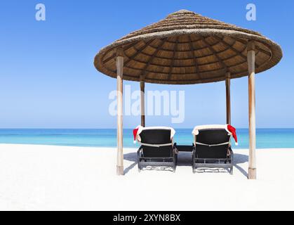 Weihnachtskarte oder Hintergrund, zwei Sonnenliegen mit Weihnachtsmützen stehen am wunderschönen tropischen Strand mit weißem Sand und türkisfarbenem Wasser. Konzept von per Stockfoto