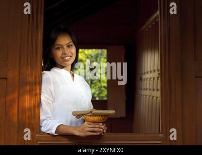 Eine attraktive Thai-Frau lächelt, während sie sich aus einem traditionellen Teakholzhaus lehnt. 20er Jahre weibliches asiatisches Thai-Modell chinesischer Abstammung Stockfoto