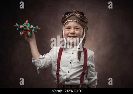 Kinderpilot mit Fliegermütze und Spielzeugflugzeug, Retro-Porträt eines Jungen in Vintage-Kleidung vor altem braunen Hintergrund Stockfoto