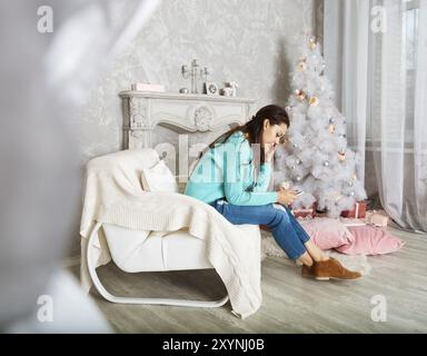 Hübsche Brünette Frau sitzt auf dem Sessel SMS auf dem Handy in der Nähe der Weihnachtsbaum Stockfoto