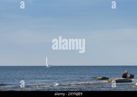 Eine einsame Jacht segelt an einem Sommertag im Abendlicht vor einer felsigen Küste. Blick auf die weißen Segel einer einsamen Yacht, die in einer hellen Sonne auf dem Meer segelt Stockfoto