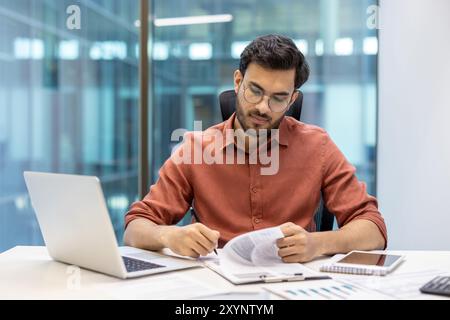 Ein seriöser Finanzier, der in einem modernen Büro sitzt, konzentriert sich auf die Prüfung von Dokumenten. Man ist umgeben von technischen Werkzeugen wie Laptop und Telefon, die eine engagierte und professionelle Arbeit veranschaulichen Stockfoto