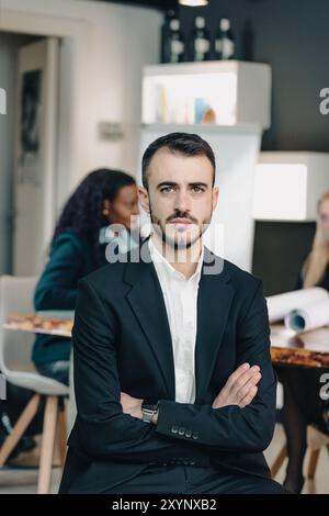 Geschäftsmann posiert mit überkreuzten Armen, während seine Geschäftspartner im Hintergrund diskutieren Stockfoto