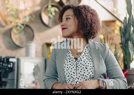 Geschäftsfrau mit lockigem Haar und Blazer sitzt in einem Café und lächelt, während sie wegblickt Stockfoto