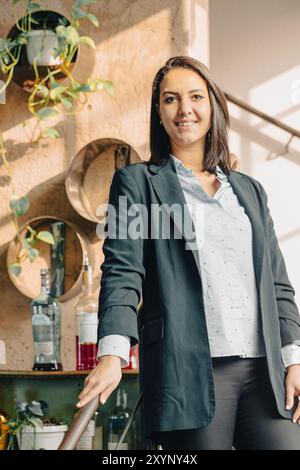 Porträt einer Geschäftsfrau, die lächelt, während sie in ihrem Büro steht, mit Pflanzen und Flaschen im Hintergrund Stockfoto