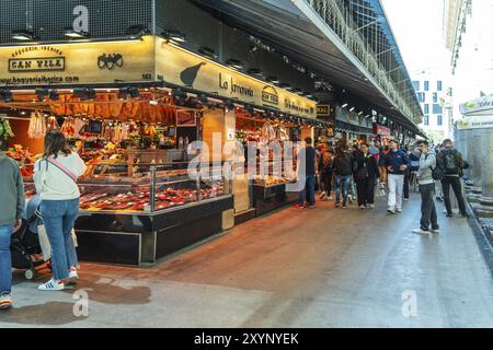 Ein beleuchteter Marktstand bietet eine reiche Auswahl an Schinken- und Fleischprodukten, Kunden im Hintergrund, Mercado de la Boqueria, berühmter Markt auf der Th Stockfoto