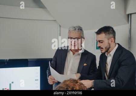 Zwei Geschäftsleute diskutieren über einige Dokumente, während sie Diagramme auf einem Bildschirm analysieren Stockfoto