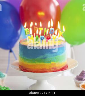 Regenbogen Kuchen und Cupcakes mit Geburtstagskerzen dekoriert. Luftballons auf dem Hintergrund Stockfoto
