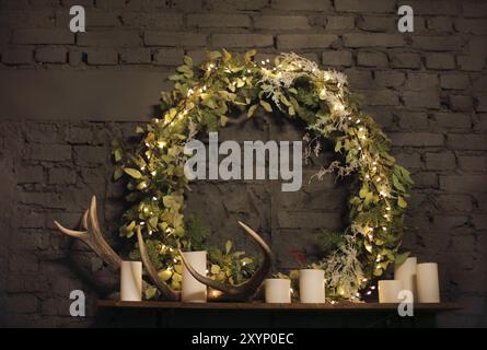 Weihnachtskranz über Mantel auf Backsteinmauerhintergrund mit Lichtern, Kerzen und Hirschgeweih Stockfoto