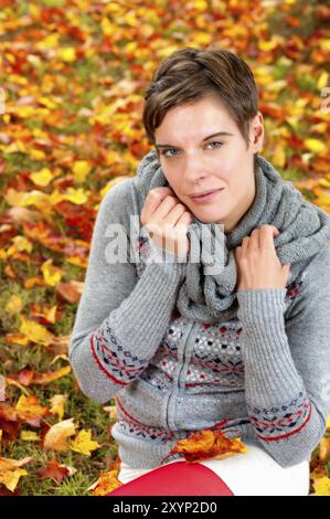 Porträt einer attraktiven jungen Frau. Die Frau sitzt in bunten Herbstblättern auf dem Boden und schaut direkt von unten in die Kamera. Hochformat Stockfoto