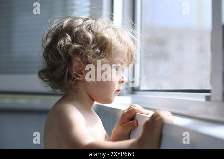 Ernsthaftes Kind schaut aufmerksam aus dem Fenster, geringe Schärfentiefe Stockfoto