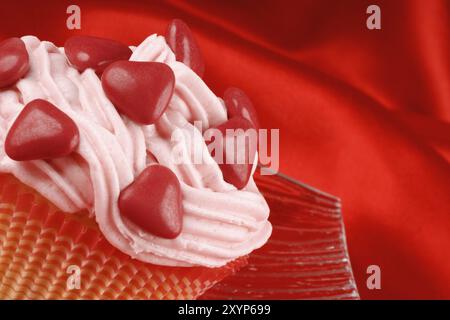 Fantasievoller Valentinstag-Cupcake, dekoriert mit kleinen roten zuckerüberzogenen Schokoladenherzen auf einer transparenten Glasschale, auf rotem Hintergrund. Selektiver lichtwellenleiter Stockfoto