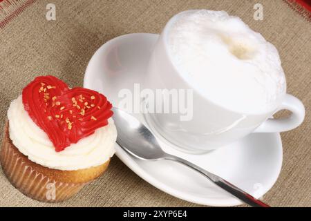 Romantisches Valentinstag-Frühstück mit Cupcake, rotem Buttercremeherz und Cappuccino vor Jute-Hintergrund. Selektiver Fokus Stockfoto