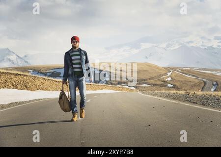 Ein stylischer bärtiger Hipster in Sonnenbrille mit einem Vintage-Rucksack spaziert an einem sonnigen Tag entlang der Asphaltstraße. Das Konzept des Anhalter- und Wanderns Stockfoto