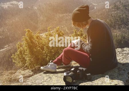 Porträt eines Hipster-Designers Fotografin mit Hut und Sonnenbrille zeichnet ein Pastellpapier in ihr Notizbuch, das draußen auf einem Felsen in den Bergen sitzt. Die Stockfoto