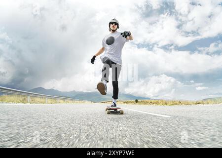 Ein Typ mit Helm und Sonnenbrille beschleunigt, indem er seinen Fuß auf seinem Longboard auf einer Landstraße auf Asphalt drückt und lächelt. Weitwinkel Stockfoto