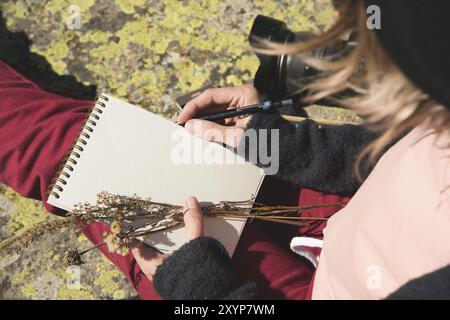 Nahaufnahme der Hände eines Mädchens, das ein leeres Notizbuch hält. Ein trockener Strauß Kräuter in der Hand und ein Bleistift. Reise-Designer-Künstler Stockfoto