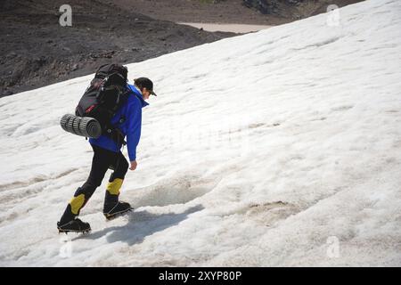 Ein Bergsteiger mit Rucksack läuft in Steigeisen entlang eines staubigen Gletschers mit Gehwegen in den Händen zwischen Rissen im Berg Stockfoto