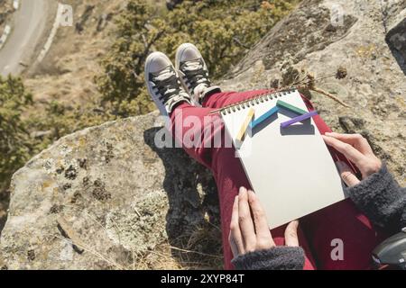 Blick von der ersten Person auf dem weiblichen Schoß ist ein Notizbuch mit Kreiden zum Zeichnen von Pastellfarben. Weibliche Hände, die ein Notizbuch in der Natur halten. Ein Mädchen sitzt Stockfoto