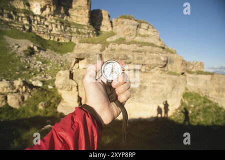 Die First-Person-Ansicht sucht mit einem Kompass in der Hand Richtung in den Sommerbergen. Suche nach Ziel. Drei Schatten von Menschen auf einem Felsen. Silhouette Stockfoto